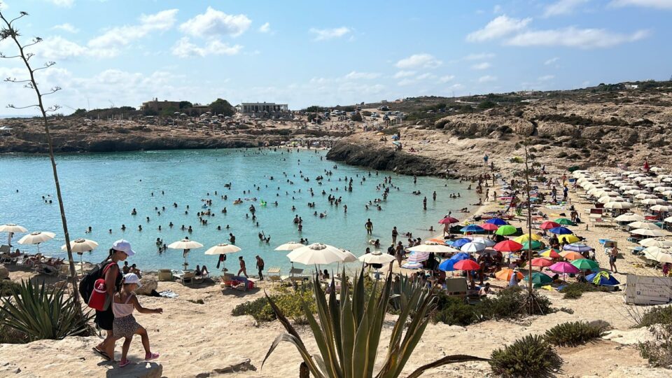 Cala Croce, una delle spiagge più amate di Lampedusa, con le sue acque turchesi e la sabbia dorata. La baia è affollata di bagnanti, con ombrelloni colorati che punteggiano la riva e le scogliere circostanti. Un'oasi perfetta per rilassarsi e godere del mare cristallino dell’isola