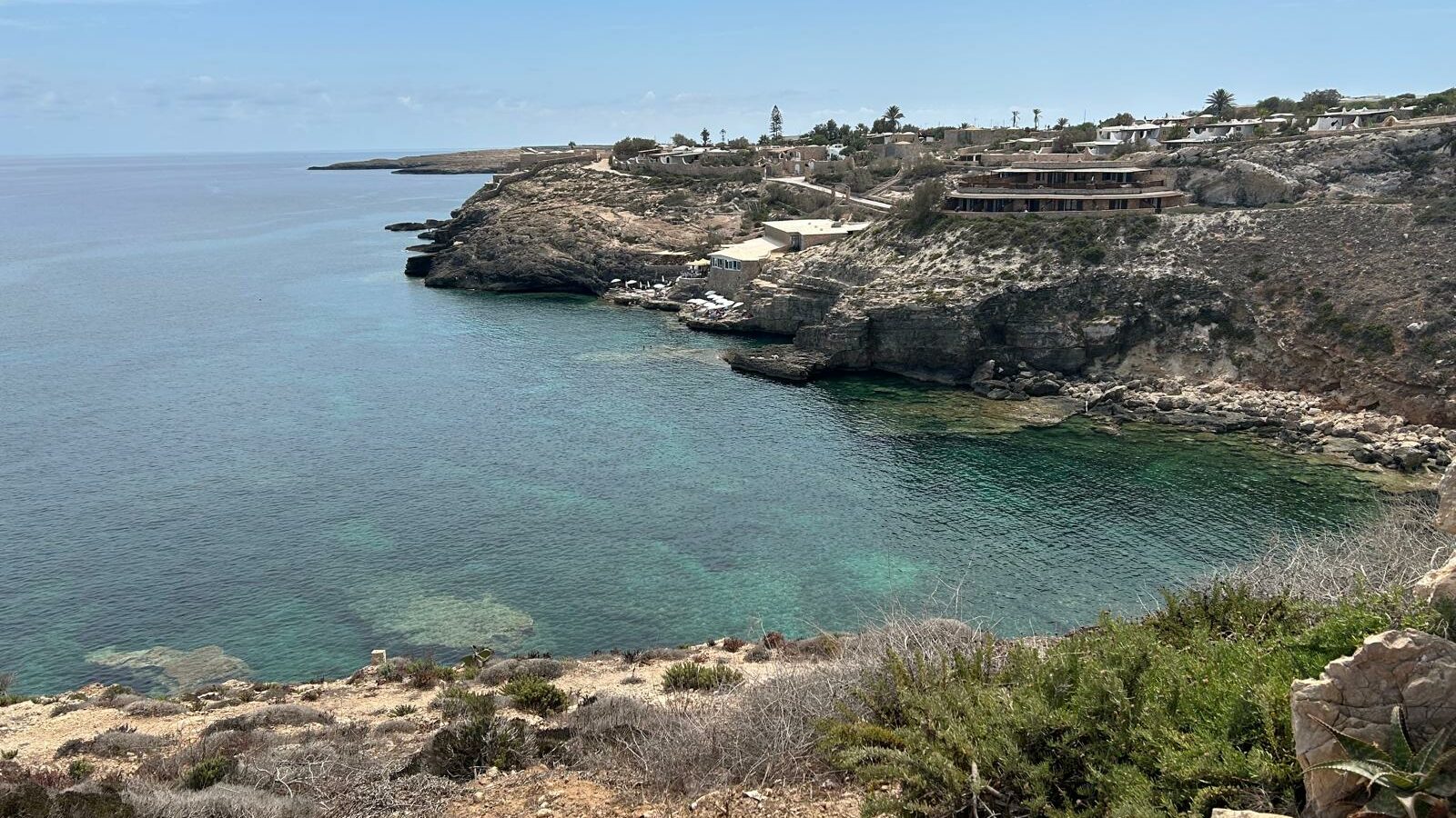 Panorama su Cala Greca, una piccola insenatura incastonata tra le scogliere di Lampedusa. Le acque trasparenti riflettono le sfumature del fondale roccioso, mentre sullo sfondo si intravedono abitazioni e strutture immerse nel paesaggio mediterraneo. Un angolo di natura incontaminata ideale per il relax e le immersioni