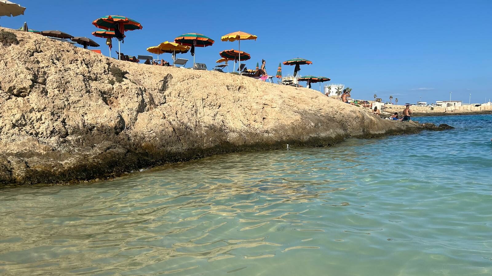Scorcio della spiaggia della Guitgia a Lampedusa, con acqua cristallina e fondale sabbioso poco profondo. Le rocce dorate ospitano colorati ombrelloni e lettini, creando un'atmosfera rilassante sotto il cielo azzurro. Sullo sfondo, bagnanti godono del sole e del mare