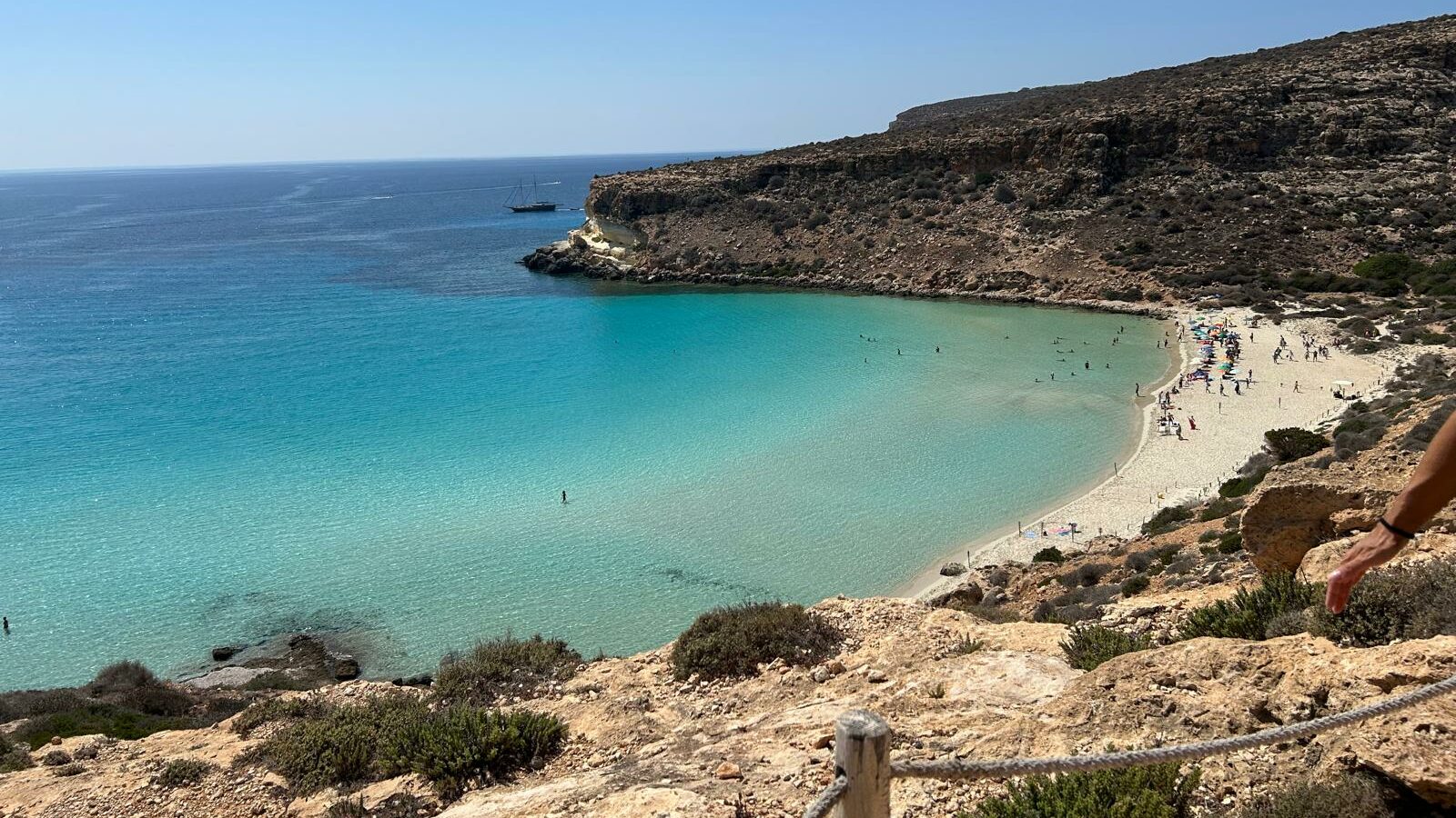 Vista panoramica della Spiaggia dei Conigli, tra le spiagge di Lampedusa la più famosa, con mare cristallino dalle sfumature turchesi, sabbia dorata e scogliere rocciose. Sullo sfondo, una barca a vela è ancorata al largo, mentre i bagnanti godono della spiaggia e dell'acqua trasparente